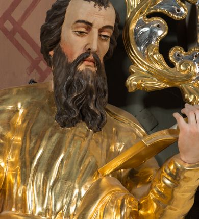 Zdjęcie nr 5: Figura pełnoplastyczna ustawiona na nieregularnej podstawie. Święty ukazany frontalnie, z pochyloną głową, lewą nogą ugiętą, opartą na podstawie. Lewą ręką podtrzymuje otwartą księgę, prawą wyciąga w bok i opiera na pile. Twarz ma pociągłą, o dojrzałych rysach, zmarszczonym czole, półprzymkniętych oczach, prostym nosie i pełnych ustach, okoloną długą, rozdwajającą się brodą i krótkimi, falowanymi włosami. Ubrany jest w złoconą tunikę, przylegającą do ciała, obficie drapowaną, przepasaną w talii oraz płaszcz zarzucony na ramiona i na lewy bok postaci. Polichromia ciała naturalistyczna, włosy brązowe. 
