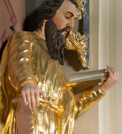 Zdjęcie nr 2: Figura pełnoplastyczna ustawiona na nieregularnej podstawie. Święty ukazany frontalnie, z pochyloną głową, lewą nogą ugiętą, opartą na podstawie. Lewą ręką podtrzymuje otwartą księgę, prawą wyciąga w bok i opiera na pile. Twarz ma pociągłą, o dojrzałych rysach, zmarszczonym czole, półprzymkniętych oczach, prostym nosie i pełnych ustach, okoloną długą, rozdwajającą się brodą i krótkimi, falowanymi włosami. Ubrany jest w złoconą tunikę, przylegającą do ciała, obficie drapowaną, przepasaną w talii oraz płaszcz zarzucony na ramiona i na lewy bok postaci. Polichromia ciała naturalistyczna, włosy brązowe. 
