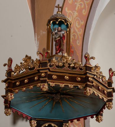 Zdjęcie nr 3: Ambona przyścienna, stojąca z dostępem w postaci jednobiegowych schodów z prawej strony, wsparta na ośmiobocznej podstawie przechodzącej w trzon zdobiony fryzem arkadkowym z kolumienkami. Kosz ośmioboczny, zamknięty profilowanym parapetem, dekorowany arkadami zamkniętymi łukiem półkolistym wspartym na skręconych kolumnach ujmujących krawędzie kosza. Wewnątrz arkad płyciny z aplikowanym motywem bukietów kwiatowych. Zaplecek w kształcie stojącego prostokąta, flankowany dwiema smukłymi kolumienkami. W polu, w płytkiej niszy dwie, rzeźbione tablice dekalogu umieszczone na obłokach. Baldachim na planie analogicznym do kosza, szerszy od niego, od dołu ujęty krenelażem, od góry rzeźbionym ornamentem ze spiętych wolut i kwiatonów, w podniebiu na jasnobłękitnym tle rzeźbiona gołębica Ducha Świętego otoczona promieniami. W zwieńczeniu figura Dobrego Pasterza pod niewielkim baldachimem, nakrytym kopułą, zakończoną krzyżem. Struktura malowana w kolorze naturalnego drewna, ornamentyka złocona, detale czerwone i srebrzone.