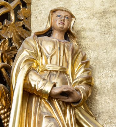 Zdjęcie nr 1: Rzeźba drążona, przedstawiająca Matkę Boską, ustawiona na niskim i półkolistym cokole. Maria zwrócona delikatnie w lewo, w kontrapoście z prawą nogą ugiętą w kolanie i z dłońmi splecionymi poniżej pasa. Twarz okrągła z wyraźnie zaznaczonym podbródkiem, małymi ustami i dużym, prostym nosem. Włosy długie, częściowo schowane pod welonem, ciemnobrązowe, opadające na plecy. Matka Boska jest ubrana w długą suknię z długimi rękawami, przewiązaną pasem oraz płaszcz, założony na głowę, na stopach ma pełne buty. Draperia szat gęsta, równolegle biegnąca w dół. Polichromia w odsłoniętych partiach ciała naturalistyczna, ubiór złocony.
