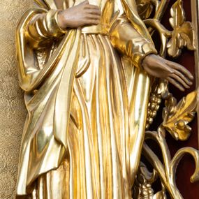 Zdjęcie nr 1: Rzeźba drążona, przedstawiająca św. Jana Ewangelistę, ustawiona na niskim, półkolistym cokole. Święty zwrócony delikatnie w prawo, w kontrapoście, z głową skierowaną w prawo i uniesioną nieco do góry, z prawą ręką złożoną na piersi, a lewą wyprostowaną i odsuniętą w bok. Twarz okrągła z wyraźnie zaznaczonym podbródkiem, małymi ustami i prostym nosem; włosy długie, ciemnobrązowe z przedziałkiem pośrodku, opadające na plecy. Święty jest ubrany w długą szatę z długimi rękawami, zapiętą w górnej części na guziki, przewiązaną pasem oraz płaszcz, którego jeden koniec trzyma w prawej dłoni. Draperia szat gęsta, równolegle biegnąca w dół. Polichromia w odsłoniętych partiach ciała naturalistyczna, szaty złocone.

