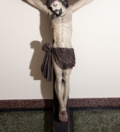 Zdjęcie nr 1: Rzeźba Chrystusa przybita do krzyża trzema gwoździami, pełna. Zbawiciel ukazany w typie Cristo vivo, w lekkim zwisie, z głową pochyloną na prawą stronę. Chrystus ma szczupłą sylwetkę ciała, delikatnie podkreśloną linię żeber. Jego twarz jest podłużna, ma długi nos, delikatnie otwarte usta i oczy, krótką brodę. Ma długie, brązowe włosy spływające na plecy i prawe ramię, a na głowie założoną koronę cierniową złożoną z dwóch gałązek. Biodra oplata brązowe perizonium, zawiązane na prawym boku z jednym końcem zwisającym wzdłuż prawego uda. Z dłoni, stóp i boku Chrystusa spływają strużki krwi. Polichromia ciała naturalistyczna, krzyż brązowy. 
