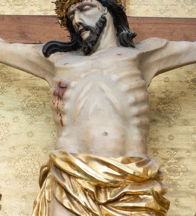Zdjęcie nr 2: Rzeźba Chrystusa przybita do krzyża trzema gwoździami. Chrystus ukazany w typie Cristo morte, w lekkim zwisie, z głową pochyloną na prawą stronę. Ma szczupłą sylwetkę ciała, podkreśloną linię żeber i mostka. Jego twarz jest podłużna, ma długi nos, otwarte usta i zamknięte oczy oraz krótką brodę. Włosy długie, ciemnobrązowe, spływają falami do przodu i na plecy. Na głowie ma założoną szeroką, złoconą koronę cierniową. Jego biodra oplata złocone perizonium, zawiązane na prawym boku ze zwisem wzdłuż prawego uda. Z dłoni, stóp i boku Chrystusa spływają strużki krwi. Nad głową wisi titulus w formie poziomej banderoli z napisem „I. N. R. I.”. Krzyż prosty, gładki, malowany na brązowo. Polichromia ciała naturalistyczna; perizonium, korona i titulus złocone.






