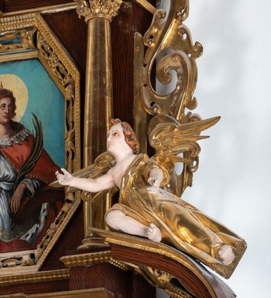 Zdjęcie nr 27: Ołtarz architektoniczny, dwukondygnacyjny, jednoosiowy ze zwieńczeniem. Mensa prostopadłościenna, antepedium w kształcie leżącego prostokąta, flankowane dwoma kanelowanymi pilastrami. W polu, w złoconej ramie dwie połączone płyciny w kształcie stojącego prostokąta wypełnione symetryczną dekoracją z akantu na błękitnym tle. Tabernakulum w formie prostopadłościennej, marmurowej skrzynki, zamknięte od góry prostym, złoconym gzymsem. Ścianka frontowa i drzwiczki obwiedzione złoconymi listwami, w polu łaciński krzyż. Cokół nastawy ustawiony na jednym stopniu, pośrodku zdobiony płyciną w kształcie leżącego prostokąta o zaokrąglonych krótszych bokach, imposty dekorowane mniejszymi płycinami wypełnionymi dekoracją roślinną. Pierwsza kondygnacja ujęta parą kolumn dźwigających przerwane belkowanie z przyczółkami, po bokach dodatkowo flankowana uszami wykończonymi ornamentem małżowinowo-chrząstkowym. W polu głównym obraz Matki Boskiej Szkaplerznej oraz św. Józefa z Dzieciątkiem i ze św. Janem Chrzcicielem na zasuwie, nad nimi para, małych uskrzydlonych aniołków. Po bokach nastawy dwie rzeźby: po prawej św. Katarzyna Aleksandryjska, a po lewej św. Barbara. Na przyczółkach usadzone dwie rzeźby aniołów z rozłożonymi szeroko rękami. Kolumny ustawione na tle prostokątnych płycin, z bazami, trzony gładkie, kapitele kompozytowe. Druga kondygnacja flankowana parą kolumn podtrzymujących wyłamane belkowanie. W polu, w ramie o kształcie stojącego prostokąta o ściętych narożach obraz św. Katarzyny Aleksandryjskiej. W zwieńczeniu promienista gloria z monogramem Marii na błękitnym tle, obwiedzionym wieńcem z obłoków. Struktura malowana w kolorze naturalnego drewna, ornamentyka złocona, detale srebrzone i błękitne.

