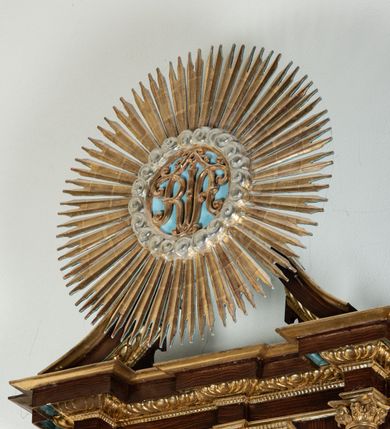 Zdjęcie nr 55: Ołtarz architektoniczny, dwukondygnacyjny, jednoosiowy ze zwieńczeniem. Mensa prostopadłościenna, antepedium w kształcie leżącego prostokąta, flankowane dwoma kanelowanymi pilastrami. W polu, w złoconej ramie dwie połączone płyciny w kształcie stojącego prostokąta wypełnione symetryczną dekoracją z akantu na błękitnym tle. Tabernakulum w formie prostopadłościennej, marmurowej skrzynki, zamknięte od góry prostym, złoconym gzymsem. Ścianka frontowa i drzwiczki obwiedzione złoconymi listwami, w polu łaciński krzyż. Cokół nastawy ustawiony na jednym stopniu, pośrodku zdobiony płyciną w kształcie leżącego prostokąta o zaokrąglonych krótszych bokach, imposty dekorowane mniejszymi płycinami wypełnionymi dekoracją roślinną. Pierwsza kondygnacja ujęta parą kolumn dźwigających przerwane belkowanie z przyczółkami, po bokach dodatkowo flankowana uszami wykończonymi ornamentem małżowinowo-chrząstkowym. W polu głównym obraz Matki Boskiej Szkaplerznej oraz św. Józefa z Dzieciątkiem i ze św. Janem Chrzcicielem na zasuwie, nad nimi para, małych uskrzydlonych aniołków. Po bokach nastawy dwie rzeźby: po prawej św. Katarzyna Aleksandryjska, a po lewej św. Barbara. Na przyczółkach usadzone dwie rzeźby aniołów z rozłożonymi szeroko rękami. Kolumny ustawione na tle prostokątnych płycin, z bazami, trzony gładkie, kapitele kompozytowe. Druga kondygnacja flankowana parą kolumn podtrzymujących wyłamane belkowanie. W polu, w ramie o kształcie stojącego prostokąta o ściętych narożach obraz św. Katarzyny Aleksandryjskiej. W zwieńczeniu promienista gloria z monogramem Marii na błękitnym tle, obwiedzionym wieńcem z obłoków. Struktura malowana w kolorze naturalnego drewna, ornamentyka złocona, detale srebrzone i błękitne.

