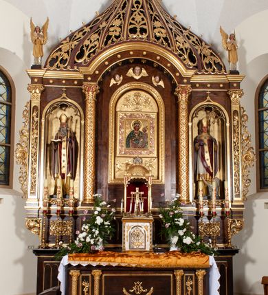 Zdjęcie nr 4: Ołtarz przyścienny, architektoniczny, trójosiowy, jednokondygnacyjny ze zwieńczeniem. Mensa prostopadłościenna. Antepedium trójosiowe, flankowane czterema kolumnami. W osi środkowej płycina w kształcie leżącego prostokąta z monogramem Matki Boskiej w polu i wieńcem z róż. W osiach bocznych płyciny w kształcie stojących prostokątów z bukietem narcyzów w polu. Tabernakulum metalowe w formie prostopadłościennej szafki zamkniętej profilowanym gzymsem wspartym na dwóch smukłych kolumnach. Drzwiczki prostokątne, zamknięte półkoliście z przedstawieniem krzyża z gorejącym sercem w czwórliściu oraz motywem winorośli i kłosów zbóż. Tron wystawienia w formie baldachimu wspartego na czterech smukłych kolumnach, zwieńczonego kopułą i rzeźbą pelikana karmiącego młode. Retabulum ustawione na wysokim cokole i predelli. Nastawa flankowana parą kolumn w części środkowej i parą pilastrów na zewnątrz. Podpory dźwigają wyłamane belkowanie, nad osią środkową wysunięte do przodu i tworzące łuk nadwieszony. W polu głównym, w ramie w kształcie stojącego prostokąta, zamkniętego łukiem półkolistym, umieszczony obraz Matki Boskiej Zembrzyckiej oraz św. Jana Chrzciciela na zasuwie. Nad polem głównym trzy uskrzydlone główki aniołków. W osiach bocznych, w niszach zamkniętych łukiem w ośli grzbiet i zwieńczonych krzyżykiem, ustawione figury świętych: po prawej św. Stanisława, po lewej św. Wojciecha. Zwieńczenie w kształcie cebulastej, ażurowej kopuły, dekorowanej ornamentem roślinnym, spiętej u góry zamkniętą koroną z krzyżykiem. Po bokach zwieńczenia, na osi pilastrów para stojących aniołów. Cokół zdobiony fryzem arkadkowym i zamknięty od góry profilowanym gzymsem. Predella pośrodku dekorowana fryzem arkadowym, a w osiach bocznych stylizowanym ornamentem roślinnym, a na skrajach zawiniętym liściem akantu. Kolumny i pilastry ustawione na bazach, kapitele kompozytowe, trzony kolumn dekorowane ukośną kratką z rozetkami w polu, a trzony pilastrów z płyciną wypełnioną wicią z róż i innych kwiatów. Po bokach ołtarz ujęty ażurowymi uszakami utworzonymi z wolutowo skręconej wici roślinnej. Fryz dekorowany rzędem rozetek. Struktura malowana w kolorze naturalnego drewna, ornamentyka i profilowania złocone, detale czerwone i bordowe, suknie aniołów srebrzone.
