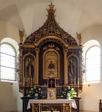 Zdjęcie nr 1: Ołtarz przyścienny, architektoniczny, trójosiowy, jednokondygnacyjny ze zwieńczeniem. Mensa prostopadłościenna. Antepedium trójosiowe, flankowane czterema kolumnami. W osi środkowej płycina w kształcie leżącego prostokąta z monogramem Matki Boskiej w polu i wieńcem z róż. W osiach bocznych płyciny w kształcie stojących prostokątów z bukietem narcyzów w polu. Tabernakulum metalowe w formie prostopadłościennej szafki zamkniętej profilowanym gzymsem wspartym na dwóch smukłych kolumnach. Drzwiczki prostokątne, zamknięte półkoliście z przedstawieniem krzyża z gorejącym sercem w czwórliściu oraz motywem winorośli i kłosów zbóż. Tron wystawienia w formie baldachimu wspartego na czterech smukłych kolumnach, zwieńczonego kopułą i rzeźbą pelikana karmiącego młode. Retabulum ustawione na wysokim cokole i predelli. Nastawa flankowana parą kolumn w części środkowej i parą pilastrów na zewnątrz. Podpory dźwigają wyłamane belkowanie, nad osią środkową wysunięte do przodu i tworzące łuk nadwieszony. W polu głównym, w ramie w kształcie stojącego prostokąta, zamkniętego łukiem półkolistym, umieszczony obraz Matki Boskiej Zembrzyckiej oraz św. Jana Chrzciciela na zasuwie. Nad polem głównym trzy uskrzydlone główki aniołków. W osiach bocznych, w niszach zamkniętych łukiem w ośli grzbiet i zwieńczonych krzyżykiem, ustawione figury świętych: po prawej św. Stanisława, po lewej św. Wojciecha. Zwieńczenie w kształcie cebulastej, ażurowej kopuły, dekorowanej ornamentem roślinnym, spiętej u góry zamkniętą koroną z krzyżykiem. Po bokach zwieńczenia, na osi pilastrów para stojących aniołów. Cokół zdobiony fryzem arkadkowym i zamknięty od góry profilowanym gzymsem. Predella pośrodku dekorowana fryzem arkadowym, a w osiach bocznych stylizowanym ornamentem roślinnym, a na skrajach zawiniętym liściem akantu. Kolumny i pilastry ustawione na bazach, kapitele kompozytowe, trzony kolumn dekorowane ukośną kratką z rozetkami w polu, a trzony pilastrów z płyciną wypełnioną wicią z róż i innych kwiatów. Po bokach ołtarz ujęty ażurowymi uszakami utworzonymi z wolutowo skręconej wici roślinnej. Fryz dekorowany rzędem rozetek. Struktura malowana w kolorze naturalnego drewna, ornamentyka i profilowania złocone, detale czerwone i bordowe, suknie aniołów srebrzone.
