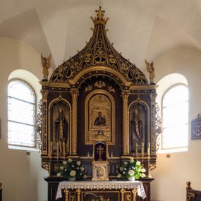 Zdjęcie nr 1: Ołtarz przyścienny, architektoniczny, trójosiowy, jednokondygnacyjny ze zwieńczeniem. Mensa prostopadłościenna. Antepedium trójosiowe, flankowane czterema kolumnami. W osi środkowej płycina w kształcie leżącego prostokąta z monogramem Matki Boskiej w polu i wieńcem z róż. W osiach bocznych płyciny w kształcie stojących prostokątów z bukietem narcyzów w polu. Tabernakulum metalowe w formie prostopadłościennej szafki zamkniętej profilowanym gzymsem wspartym na dwóch smukłych kolumnach. Drzwiczki prostokątne, zamknięte półkoliście z przedstawieniem krzyża z gorejącym sercem w czwórliściu oraz motywem winorośli i kłosów zbóż. Tron wystawienia w formie baldachimu wspartego na czterech smukłych kolumnach, zwieńczonego kopułą i rzeźbą pelikana karmiącego młode. Retabulum ustawione na wysokim cokole i predelli. Nastawa flankowana parą kolumn w części środkowej i parą pilastrów na zewnątrz. Podpory dźwigają wyłamane belkowanie, nad osią środkową wysunięte do przodu i tworzące łuk nadwieszony. W polu głównym, w ramie w kształcie stojącego prostokąta, zamkniętego łukiem półkolistym, umieszczony obraz Matki Boskiej Zembrzyckiej oraz św. Jana Chrzciciela na zasuwie. Nad polem głównym trzy uskrzydlone główki aniołków. W osiach bocznych, w niszach zamkniętych łukiem w ośli grzbiet i zwieńczonych krzyżykiem, ustawione figury świętych: po prawej św. Stanisława, po lewej św. Wojciecha. Zwieńczenie w kształcie cebulastej, ażurowej kopuły, dekorowanej ornamentem roślinnym, spiętej u góry zamkniętą koroną z krzyżykiem. Po bokach zwieńczenia, na osi pilastrów para stojących aniołów. Cokół zdobiony fryzem arkadkowym i zamknięty od góry profilowanym gzymsem. Predella pośrodku dekorowana fryzem arkadowym, a w osiach bocznych stylizowanym ornamentem roślinnym, a na skrajach zawiniętym liściem akantu. Kolumny i pilastry ustawione na bazach, kapitele kompozytowe, trzony kolumn dekorowane ukośną kratką z rozetkami w polu, a trzony pilastrów z płyciną wypełnioną wicią z róż i innych kwiatów. Po bokach ołtarz ujęty ażurowymi uszakami utworzonymi z wolutowo skręconej wici roślinnej. Fryz dekorowany rzędem rozetek. Struktura malowana w kolorze naturalnego drewna, ornamentyka i profilowania złocone, detale czerwone i bordowe, suknie aniołów srebrzone.
