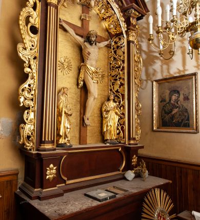 Zdjęcie nr 3: Nastawa ołtarzowa architektoniczna, jednokondygnacyjna i jednoosiowa. Ołtarz prostopadłościenny, mensa drewniana z kamiennym portatylem. Cokół nastawy ustawiony na ołtarzu pośrodku zdobiony płyciną w kształcie leżącego prostokąta o zaokrąglonych krótszych bokach, imposty dekorowane rozetkami. Kondygnacja ujęta parą pięciobocznych pilastrów dźwigających przerwane belkowanie z przyczółkami, po bokach dodatkowo flankowana uszami wykończonymi ornamentem małżowinowo-chrząstkowym. Pilastry ustawione na bazach, mają kanelowane trzony i kompozytowe kapitele. W polu głównym w głębokiej wnęce umieszczony jest krucyfiks oraz rzeźba Matki Boskiej po lewej stronie i św. Jana Ewangelisty po prawej. Tło wyłożono tkaniną. Po bokach krucyfiksu zawieszone są dwa gorejące serca na tle promienistej glorii. Obramienie wnęki wykończone ażurową wicią winorośli po bokach, a w łuku podwieszonymi szyszkami. Nad wnęką uskrzydlona główka aniołka z szyją podwiązaną chustą, skierowana na wprost. Analogiczne rzeźby znajdują się w partii fryzu na osi kolumn. Na przyczółkach ułożona jest para aniołków z arma Christi w dłoniach. W zwieńczeniu promienista gloria z monogramem Chrystusa na czerwonym tle, obwiedzionym wieńcem z obłoków. Struktura malowana w kolorze naturalnego drewna, ornamentyka złocona, detale czerwone.
