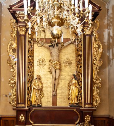 Zdjęcie nr 1: Nastawa ołtarzowa architektoniczna, jednokondygnacyjna i jednoosiowa. Ołtarz prostopadłościenny, mensa drewniana z kamiennym portatylem. Cokół nastawy ustawiony na ołtarzu pośrodku zdobiony płyciną w kształcie leżącego prostokąta o zaokrąglonych krótszych bokach, imposty dekorowane rozetkami. Kondygnacja ujęta parą pięciobocznych pilastrów dźwigających przerwane belkowanie z przyczółkami, po bokach dodatkowo flankowana uszami wykończonymi ornamentem małżowinowo-chrząstkowym. Pilastry ustawione na bazach, mają kanelowane trzony i kompozytowe kapitele. W polu głównym w głębokiej wnęce umieszczony jest krucyfiks oraz rzeźba Matki Boskiej po lewej stronie i św. Jana Ewangelisty po prawej. Tło wyłożono tkaniną. Po bokach krucyfiksu zawieszone są dwa gorejące serca na tle promienistej glorii. Obramienie wnęki wykończone ażurową wicią winorośli po bokach, a w łuku podwieszonymi szyszkami. Nad wnęką uskrzydlona główka aniołka z szyją podwiązaną chustą, skierowana na wprost. Analogiczne rzeźby znajdują się w partii fryzu na osi kolumn. Na przyczółkach ułożona jest para aniołków z arma Christi w dłoniach. W zwieńczeniu promienista gloria z monogramem Chrystusa na czerwonym tle, obwiedzionym wieńcem z obłoków. Struktura malowana w kolorze naturalnego drewna, ornamentyka złocona, detale czerwone.
