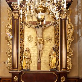 Zdjęcie nr 1: Nastawa ołtarzowa architektoniczna, jednokondygnacyjna i jednoosiowa. Ołtarz prostopadłościenny, mensa drewniana z kamiennym portatylem. Cokół nastawy ustawiony na ołtarzu pośrodku zdobiony płyciną w kształcie leżącego prostokąta o zaokrąglonych krótszych bokach, imposty dekorowane rozetkami. Kondygnacja ujęta parą pięciobocznych pilastrów dźwigających przerwane belkowanie z przyczółkami, po bokach dodatkowo flankowana uszami wykończonymi ornamentem małżowinowo-chrząstkowym. Pilastry ustawione na bazach, mają kanelowane trzony i kompozytowe kapitele. W polu głównym w głębokiej wnęce umieszczony jest krucyfiks oraz rzeźba Matki Boskiej po lewej stronie i św. Jana Ewangelisty po prawej. Tło wyłożono tkaniną. Po bokach krucyfiksu zawieszone są dwa gorejące serca na tle promienistej glorii. Obramienie wnęki wykończone ażurową wicią winorośli po bokach, a w łuku podwieszonymi szyszkami. Nad wnęką uskrzydlona główka aniołka z szyją podwiązaną chustą, skierowana na wprost. Analogiczne rzeźby znajdują się w partii fryzu na osi kolumn. Na przyczółkach ułożona jest para aniołków z arma Christi w dłoniach. W zwieńczeniu promienista gloria z monogramem Chrystusa na czerwonym tle, obwiedzionym wieńcem z obłoków. Struktura malowana w kolorze naturalnego drewna, ornamentyka złocona, detale czerwone.
