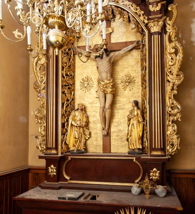 Zdjęcie nr 4: Nastawa ołtarzowa architektoniczna, jednokondygnacyjna i jednoosiowa. Ołtarz prostopadłościenny, mensa drewniana z kamiennym portatylem. Cokół nastawy ustawiony na ołtarzu pośrodku zdobiony płyciną w kształcie leżącego prostokąta o zaokrąglonych krótszych bokach, imposty dekorowane rozetkami. Kondygnacja ujęta parą pięciobocznych pilastrów dźwigających przerwane belkowanie z przyczółkami, po bokach dodatkowo flankowana uszami wykończonymi ornamentem małżowinowo-chrząstkowym. Pilastry ustawione na bazach, mają kanelowane trzony i kompozytowe kapitele. W polu głównym w głębokiej wnęce umieszczony jest krucyfiks oraz rzeźba Matki Boskiej po lewej stronie i św. Jana Ewangelisty po prawej. Tło wyłożono tkaniną. Po bokach krucyfiksu zawieszone są dwa gorejące serca na tle promienistej glorii. Obramienie wnęki wykończone ażurową wicią winorośli po bokach, a w łuku podwieszonymi szyszkami. Nad wnęką uskrzydlona główka aniołka z szyją podwiązaną chustą, skierowana na wprost. Analogiczne rzeźby znajdują się w partii fryzu na osi kolumn. Na przyczółkach ułożona jest para aniołków z arma Christi w dłoniach. W zwieńczeniu promienista gloria z monogramem Chrystusa na czerwonym tle, obwiedzionym wieńcem z obłoków. Struktura malowana w kolorze naturalnego drewna, ornamentyka złocona, detale czerwone.
