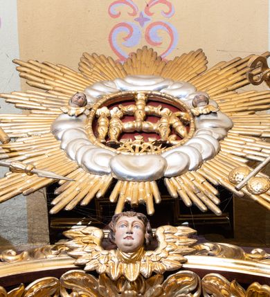 Zdjęcie nr 5: Nastawa ołtarzowa architektoniczna, jednokondygnacyjna i jednoosiowa. Ołtarz prostopadłościenny, mensa drewniana z kamiennym portatylem. Cokół nastawy ustawiony na ołtarzu pośrodku zdobiony płyciną w kształcie leżącego prostokąta o zaokrąglonych krótszych bokach, imposty dekorowane rozetkami. Kondygnacja ujęta parą pięciobocznych pilastrów dźwigających przerwane belkowanie z przyczółkami, po bokach dodatkowo flankowana uszami wykończonymi ornamentem małżowinowo-chrząstkowym. Pilastry ustawione na bazach, mają kanelowane trzony i kompozytowe kapitele. W polu głównym w głębokiej wnęce umieszczony jest krucyfiks oraz rzeźba Matki Boskiej po lewej stronie i św. Jana Ewangelisty po prawej. Tło wyłożono tkaniną. Po bokach krucyfiksu zawieszone są dwa gorejące serca na tle promienistej glorii. Obramienie wnęki wykończone ażurową wicią winorośli po bokach, a w łuku podwieszonymi szyszkami. Nad wnęką uskrzydlona główka aniołka z szyją podwiązaną chustą, skierowana na wprost. Analogiczne rzeźby znajdują się w partii fryzu na osi kolumn. Na przyczółkach ułożona jest para aniołków z arma Christi w dłoniach. W zwieńczeniu promienista gloria z monogramem Chrystusa na czerwonym tle, obwiedzionym wieńcem z obłoków. Struktura malowana w kolorze naturalnego drewna, ornamentyka złocona, detale czerwone.
