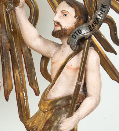 Zdjęcie nr 6: Pokrywa chrzcielnicy na planie koła z dekoracją imitującą puklowanie. Na szczycie pokrywy umieszczona rzeźbiona scena przedstawiająca Chrzest Chrystusa w Jordanie. Po lewej stronie Jezus stopami zanurzony w wodach Jordanu, zwrócony trzy czwarte w stronę św. Jana Chrzciciela, z rękami złożonymi na piersiach w geście pokory. Twarz podłużna, okolona brodą z długim nosem i wzrokiem skierowanym w dół. Włosy długie, opadające na plecy. Ubrany jest w złocone perizonium, luźno zawiązane na biodrach. Po prawej św. Jan Chrzciciel klęczący na kamiennym postumencie, zwrócony w prawo, w lewej ręce trzymający krzyż, prawą polewający wodą Chrystusa. Twarz podłużna, okolona krótką brodą i długimi włosami. Ubrany jest w skórę wielbłądzią, odsłaniającą nagi tors i nogi. Krzyż długi z napisem na banderoli: „OTO BARANEK BOŻY”. Za nimi zatknięta jest duża, promienista gloria, pośrodku której znajduje się gołębica Ducha Świętego, a nad nią półpostać Boga Ojca wynurzająca się z obłoków z rozłożonymi szeroko rękami. Bóg Ojciec o rysach twarzy starszego mężczyzny z siwymi włosami i brodą, ubrany w złoconą suknię. Polichromia w odsłoniętych partiach ciała naturalistyczna, podstawa pokrywy, gloria i szaty złocone, obłoki srebrzone.


