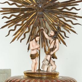 Zdjęcie nr 1: Pokrywa chrzcielnicy na planie koła z dekoracją imitującą puklowanie. Na szczycie pokrywy umieszczona rzeźbiona scena przedstawiająca Chrzest Chrystusa w Jordanie. Po lewej stronie Jezus stopami zanurzony w wodach Jordanu, zwrócony trzy czwarte w stronę św. Jana Chrzciciela, z rękami złożonymi na piersiach w geście pokory. Twarz podłużna, okolona brodą z długim nosem i wzrokiem skierowanym w dół. Włosy długie, opadające na plecy. Ubrany jest w złocone perizonium, luźno zawiązane na biodrach. Po prawej św. Jan Chrzciciel klęczący na kamiennym postumencie, zwrócony w prawo, w lewej ręce trzymający krzyż, prawą polewający wodą Chrystusa. Twarz podłużna, okolona krótką brodą i długimi włosami. Ubrany jest w skórę wielbłądzią, odsłaniającą nagi tors i nogi. Krzyż długi z napisem na banderoli: „OTO BARANEK BOŻY”. Za nimi zatknięta jest duża, promienista gloria, pośrodku której znajduje się gołębica Ducha Świętego, a nad nią półpostać Boga Ojca wynurzająca się z obłoków z rozłożonymi szeroko rękami. Bóg Ojciec o rysach twarzy starszego mężczyzny z siwymi włosami i brodą, ubrany w złoconą suknię. Polichromia w odsłoniętych partiach ciała naturalistyczna, podstawa pokrywy, gloria i szaty złocone, obłoki srebrzone.



