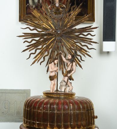 Zdjęcie nr 2: Pokrywa chrzcielnicy na planie koła z dekoracją imitującą puklowanie. Na szczycie pokrywy umieszczona rzeźbiona scena przedstawiająca Chrzest Chrystusa w Jordanie. Po lewej stronie Jezus stopami zanurzony w wodach Jordanu, zwrócony trzy czwarte w stronę św. Jana Chrzciciela, z rękami złożonymi na piersiach w geście pokory. Twarz podłużna, okolona brodą z długim nosem i wzrokiem skierowanym w dół. Włosy długie, opadające na plecy. Ubrany jest w złocone perizonium, luźno zawiązane na biodrach. Po prawej św. Jan Chrzciciel klęczący na kamiennym postumencie, zwrócony w prawo, w lewej ręce trzymający krzyż, prawą polewający wodą Chrystusa. Twarz podłużna, okolona krótką brodą i długimi włosami. Ubrany jest w skórę wielbłądzią, odsłaniającą nagi tors i nogi. Krzyż długi z napisem na banderoli: „OTO BARANEK BOŻY”. Za nimi zatknięta jest duża, promienista gloria, pośrodku której znajduje się gołębica Ducha Świętego, a nad nią półpostać Boga Ojca wynurzająca się z obłoków z rozłożonymi szeroko rękami. Bóg Ojciec o rysach twarzy starszego mężczyzny z siwymi włosami i brodą, ubrany w złoconą suknię. Polichromia w odsłoniętych partiach ciała naturalistyczna, podstawa pokrywy, gloria i szaty złocone, obłoki srebrzone.


