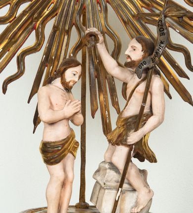 Zdjęcie nr 5: Pokrywa chrzcielnicy na planie koła z dekoracją imitującą puklowanie. Na szczycie pokrywy umieszczona rzeźbiona scena przedstawiająca Chrzest Chrystusa w Jordanie. Po lewej stronie Jezus stopami zanurzony w wodach Jordanu, zwrócony trzy czwarte w stronę św. Jana Chrzciciela, z rękami złożonymi na piersiach w geście pokory. Twarz podłużna, okolona brodą z długim nosem i wzrokiem skierowanym w dół. Włosy długie, opadające na plecy. Ubrany jest w złocone perizonium, luźno zawiązane na biodrach. Po prawej św. Jan Chrzciciel klęczący na kamiennym postumencie, zwrócony w prawo, w lewej ręce trzymający krzyż, prawą polewający wodą Chrystusa. Twarz podłużna, okolona krótką brodą i długimi włosami. Ubrany jest w skórę wielbłądzią, odsłaniającą nagi tors i nogi. Krzyż długi z napisem na banderoli: „OTO BARANEK BOŻY”. Za nimi zatknięta jest duża, promienista gloria, pośrodku której znajduje się gołębica Ducha Świętego, a nad nią półpostać Boga Ojca wynurzająca się z obłoków z rozłożonymi szeroko rękami. Bóg Ojciec o rysach twarzy starszego mężczyzny z siwymi włosami i brodą, ubrany w złoconą suknię. Polichromia w odsłoniętych partiach ciała naturalistyczna, podstawa pokrywy, gloria i szaty złocone, obłoki srebrzone.


