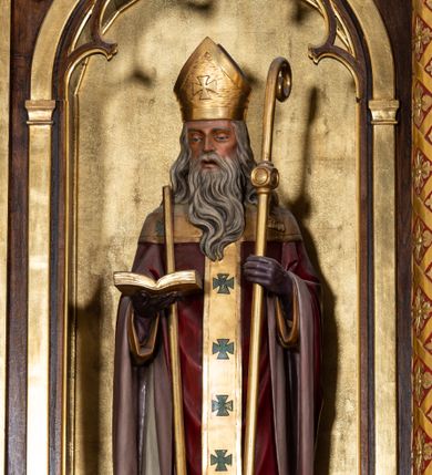 Zdjęcie nr 1: Rzeźba przedstawia św. Wojciecha, ukazanego frontalnie w postawie stojącej. Święty w lewej ręce trzyma pastorał, a w prawej otwartą księgę, o prawe ramię ma oparte wiosło. Twarz szczupła o wyraźnie wystających kościach policzkowych, otwartych ustach, krótkim nosie i wzroku skierowanym na wprost. Głowę okalają długie, siwe i falowane włosy oraz długa broda. Święty jest ubrany w żółtą albę, białą komżę zdobioną u dołu złotym pasem, bordowy ornat i rękawice, złoty paliusz oraz złotą infułę na głowie. Rzeźba ustawiona jest na niskim, brązowym, prostopadłościennym cokole. Polichromia w odsłoniętych partiach ciała naturalistyczna, atrybuty i detale złocone.

