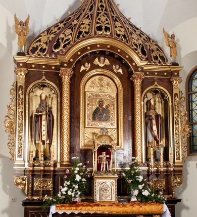 Zdjęcie nr 3: Rzeźba przedstawia św. Stanisława, ukazanego frontalnie w postawie stojącej. Święty w lewej ręce trzyma pastorał, a prawą ugiętą w łokciu wyciąga przed siebie. Twarz owalna o dużych ustach i nosie, wzroku skierowanym na wprost. Głowę okalają średniej długości włosy, silnie skręcone, zasłaniające uszy. Święty jest ubrany w żółtą albę, białą komżę zdobioną u dołu złotym pasem, bordowy ornat i rękawice, złoty paliusz oraz złotą infułę na głowie. U dołu po prawej stronie widoczny jest w półpostaci i z lewego profilu Piotrowin z lewą ręką uniesioną do góry. Ubrany jest w białą tkaninę nałożoną na głowę. Rzeźby ustawione są na niskim, brązowym, prostopadłościennym cokole. Polichromia w odsłoniętych partiach ciała naturalistyczna, atrybuty i detale złocone.
