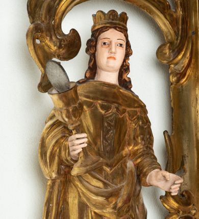 Zdjęcie nr 4: Rzeźba przedstawiająca św. Barbarę, ustawiona na niskim cokole. Święta ukazana frontalnie w delikatnym kontrapoście, z kielichem z hostią w prawej dłoni oraz lewą ręką ugiętą w łokciu i wyciągniętą przed siebie. Twarz owalna z małymi ustami, długim i szerokim nosem, migdałowymi oczami. Włosy długie, brązowe, opadające falami na plecy. Święta ubrana jest w złotą, długą suknię z długimi rękawami oraz złoty płaszcz, na stopach ma założone buty, a na głowę otwartą koronę. Po jej lewej stronie ustawiona jest miniaturowa, dwukondygnacyjna wieża z prostokątnym otworem drzwiowym i trzema okrągłymi oknami, zamknięta kopułą. Kielich na okrągłej stopie, z trzonem przechodzącym w wąską tuleję, oddzieloną od czary półkolistym talerzykiem, czara w dolnej części zdobiona puklowaniem. Polichromia w odsłoniętych partiach ciała naturalistyczna, szaty i atrybuty złocone i srebrzone, detale błękitne (wnętrze korony i monogram na hostii).





