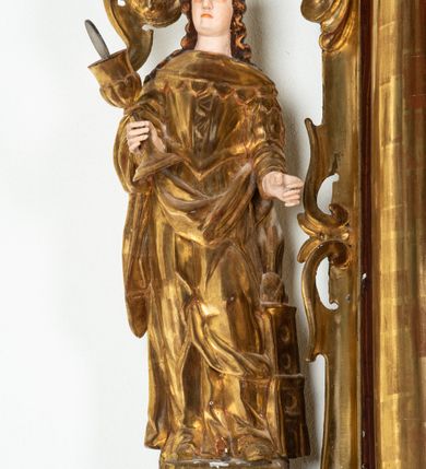 Zdjęcie nr 1: Rzeźba przedstawiająca św. Barbarę, ustawiona na niskim cokole. Święta ukazana frontalnie w delikatnym kontrapoście, z kielichem z hostią w prawej dłoni oraz lewą ręką ugiętą w łokciu i wyciągniętą przed siebie. Twarz owalna z małymi ustami, długim i szerokim nosem, migdałowymi oczami. Włosy długie, brązowe, opadające falami na plecy. Święta ubrana jest w złotą, długą suknię z długimi rękawami oraz złoty płaszcz, na stopach ma założone buty, a na głowę otwartą koronę. Po jej lewej stronie ustawiona jest miniaturowa, dwukondygnacyjna wieża z prostokątnym otworem drzwiowym i trzema okrągłymi oknami, zamknięta kopułą. Kielich na okrągłej stopie, z trzonem przechodzącym w wąską tuleję, oddzieloną od czary półkolistym talerzykiem, czara w dolnej części zdobiona puklowaniem. Polichromia w odsłoniętych partiach ciała naturalistyczna, szaty i atrybuty złocone i srebrzone, detale błękitne (wnętrze korony i monogram na hostii).




