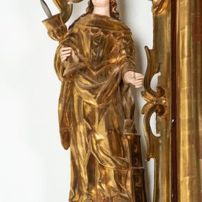 Zdjęcie nr 1: Rzeźba przedstawiająca św. Barbarę, ustawiona na niskim cokole. Święta ukazana frontalnie w delikatnym kontrapoście, z kielichem z hostią w prawej dłoni oraz lewą ręką ugiętą w łokciu i wyciągniętą przed siebie. Twarz owalna z małymi ustami, długim i szerokim nosem, migdałowymi oczami. Włosy długie, brązowe, opadające falami na plecy. Święta ubrana jest w złotą, długą suknię z długimi rękawami oraz złoty płaszcz, na stopach ma założone buty, a na głowę otwartą koronę. Po jej lewej stronie ustawiona jest miniaturowa, dwukondygnacyjna wieża z prostokątnym otworem drzwiowym i trzema okrągłymi oknami, zamknięta kopułą. Kielich na okrągłej stopie, z trzonem przechodzącym w wąską tuleję, oddzieloną od czary półkolistym talerzykiem, czara w dolnej części zdobiona puklowaniem. Polichromia w odsłoniętych partiach ciała naturalistyczna, szaty i atrybuty złocone i srebrzone, detale błękitne (wnętrze korony i monogram na hostii).




