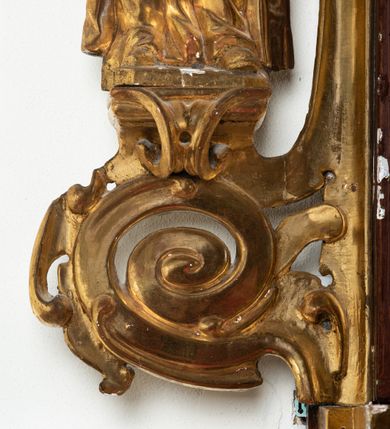 Zdjęcie nr 7: Rzeźba przedstawiająca św. Barbarę, ustawiona na niskim cokole. Święta ukazana frontalnie w delikatnym kontrapoście, z kielichem z hostią w prawej dłoni oraz lewą ręką ugiętą w łokciu i wyciągniętą przed siebie. Twarz owalna z małymi ustami, długim i szerokim nosem, migdałowymi oczami. Włosy długie, brązowe, opadające falami na plecy. Święta ubrana jest w złotą, długą suknię z długimi rękawami oraz złoty płaszcz, na stopach ma założone buty, a na głowę otwartą koronę. Po jej lewej stronie ustawiona jest miniaturowa, dwukondygnacyjna wieża z prostokątnym otworem drzwiowym i trzema okrągłymi oknami, zamknięta kopułą. Kielich na okrągłej stopie, z trzonem przechodzącym w wąską tuleję, oddzieloną od czary półkolistym talerzykiem, czara w dolnej części zdobiona puklowaniem. Polichromia w odsłoniętych partiach ciała naturalistyczna, szaty i atrybuty złocone i srebrzone, detale błękitne (wnętrze korony i monogram na hostii).




