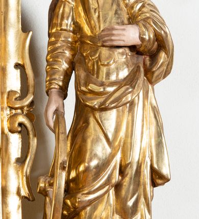 Zdjęcie nr 3: Rzeźba przedstawiająca św. Katarzynę Aleksandryjską, ustawiona na niskim cokole. Święta ukazana frontalnie w delikatnym kontrapoście, z sylwetką przechyloną w prawą stronę, z lewą ręką złożoną na tułowiu, a prawą opartą o złamane koło. Twarz owalna z małymi ustami, długim i wąskim nosem, migdałowymi oczami. Włosy długie, brązowe, opadające falami na plecy i wzdłuż ramion. Święta ubrana jest w złotą, długą suknię z długimi rękawami oraz złoty płaszcz, na stopach ma założone buty, a na głowę otwartą koronę. Polichromia w odsłoniętych partiach ciała naturalistyczna, szaty i atrybuty złocone i srebrzone, detale błękitne (wnętrze korony).



