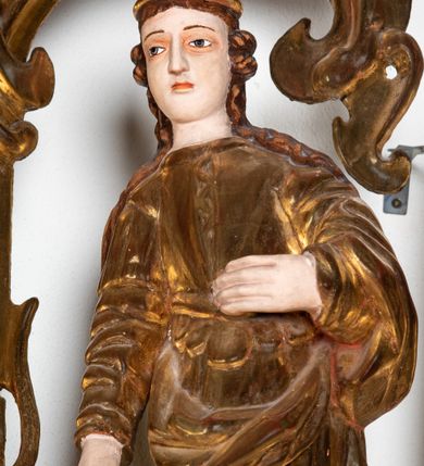 Zdjęcie nr 7: Rzeźba przedstawiająca św. Katarzynę Aleksandryjską, ustawiona na niskim cokole. Święta ukazana frontalnie w delikatnym kontrapoście, z sylwetką przechyloną w prawą stronę, z lewą ręką złożoną na tułowiu, a prawą opartą o złamane koło. Twarz owalna z małymi ustami, długim i wąskim nosem, migdałowymi oczami. Włosy długie, brązowe, opadające falami na plecy i wzdłuż ramion. Święta ubrana jest w złotą, długą suknię z długimi rękawami oraz złoty płaszcz, na stopach ma założone buty, a na głowę otwartą koronę. Polichromia w odsłoniętych partiach ciała naturalistyczna, szaty i atrybuty złocone i srebrzone, detale błękitne (wnętrze korony).



