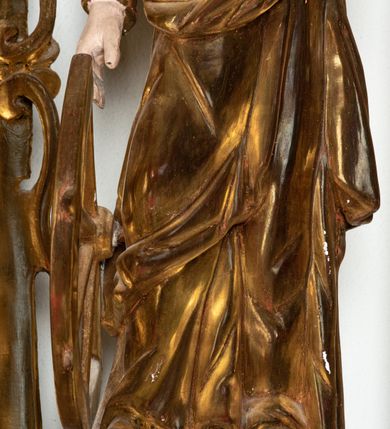 Zdjęcie nr 8: Rzeźba przedstawiająca św. Katarzynę Aleksandryjską, ustawiona na niskim cokole. Święta ukazana frontalnie w delikatnym kontrapoście, z sylwetką przechyloną w prawą stronę, z lewą ręką złożoną na tułowiu, a prawą opartą o złamane koło. Twarz owalna z małymi ustami, długim i wąskim nosem, migdałowymi oczami. Włosy długie, brązowe, opadające falami na plecy i wzdłuż ramion. Święta ubrana jest w złotą, długą suknię z długimi rękawami oraz złoty płaszcz, na stopach ma założone buty, a na głowę otwartą koronę. Polichromia w odsłoniętych partiach ciała naturalistyczna, szaty i atrybuty złocone i srebrzone, detale błękitne (wnętrze korony).



