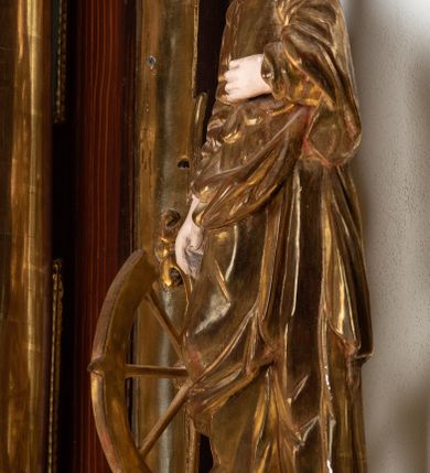 Zdjęcie nr 4: Rzeźba przedstawiająca św. Katarzynę Aleksandryjską, ustawiona na niskim cokole. Święta ukazana frontalnie w delikatnym kontrapoście, z sylwetką przechyloną w prawą stronę, z lewą ręką złożoną na tułowiu, a prawą opartą o złamane koło. Twarz owalna z małymi ustami, długim i wąskim nosem, migdałowymi oczami. Włosy długie, brązowe, opadające falami na plecy i wzdłuż ramion. Święta ubrana jest w złotą, długą suknię z długimi rękawami oraz złoty płaszcz, na stopach ma założone buty, a na głowę otwartą koronę. Polichromia w odsłoniętych partiach ciała naturalistyczna, szaty i atrybuty złocone i srebrzone, detale błękitne (wnętrze korony).



