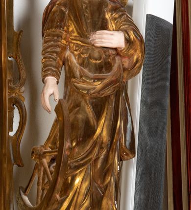 Zdjęcie nr 6: Rzeźba przedstawiająca św. Katarzynę Aleksandryjską, ustawiona na niskim cokole. Święta ukazana frontalnie w delikatnym kontrapoście, z sylwetką przechyloną w prawą stronę, z lewą ręką złożoną na tułowiu, a prawą opartą o złamane koło. Twarz owalna z małymi ustami, długim i wąskim nosem, migdałowymi oczami. Włosy długie, brązowe, opadające falami na plecy i wzdłuż ramion. Święta ubrana jest w złotą, długą suknię z długimi rękawami oraz złoty płaszcz, na stopach ma założone buty, a na głowę otwartą koronę. Polichromia w odsłoniętych partiach ciała naturalistyczna, szaty i atrybuty złocone i srebrzone, detale błękitne (wnętrze korony).



