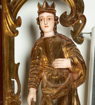 Zdjęcie nr 1: Rzeźba przedstawiająca św. Katarzynę Aleksandryjską, ustawiona na niskim cokole. Święta ukazana frontalnie w delikatnym kontrapoście, z sylwetką przechyloną w prawą stronę, z lewą ręką złożoną na tułowiu, a prawą opartą o złamane koło. Twarz owalna z małymi ustami, długim i wąskim nosem, migdałowymi oczami. Włosy długie, brązowe, opadające falami na plecy i wzdłuż ramion. Święta ubrana jest w złotą, długą suknię z długimi rękawami oraz złoty płaszcz, na stopach ma założone buty, a na głowę otwartą koronę. Polichromia w odsłoniętych partiach ciała naturalistyczna, szaty i atrybuty złocone i srebrzone, detale błękitne (wnętrze korony).



