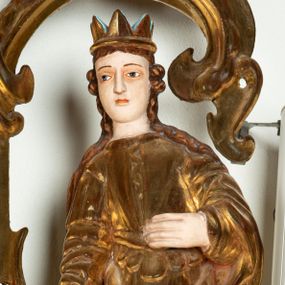 Zdjęcie nr 1: Rzeźba przedstawiająca św. Katarzynę Aleksandryjską, ustawiona na niskim cokole. Święta ukazana frontalnie w delikatnym kontrapoście, z sylwetką przechyloną w prawą stronę, z lewą ręką złożoną na tułowiu, a prawą opartą o złamane koło. Twarz owalna z małymi ustami, długim i wąskim nosem, migdałowymi oczami. Włosy długie, brązowe, opadające falami na plecy i wzdłuż ramion. Święta ubrana jest w złotą, długą suknię z długimi rękawami oraz złoty płaszcz, na stopach ma założone buty, a na głowę otwartą koronę. Polichromia w odsłoniętych partiach ciała naturalistyczna, szaty i atrybuty złocone i srebrzone, detale błękitne (wnętrze korony).



