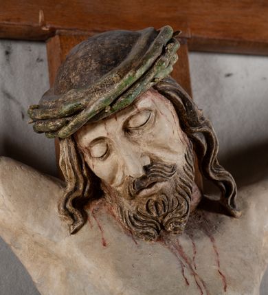 Zdjęcie nr 3: Rzeźba Chrystusa przybita do krzyża trzema gwoździami, pełna. Zbawiciel ukazany w typie Cristo morte, w lekkim zwisie, z głową pochyloną na prawą stronę. Jezus ma szczupłą sylwetkę ciała, duże stopy, delikatnie podkreśloną linię żeber. Jego twarz jest podłużna, ma długi nos, delikatnie otwarte usta i zamknięte oczy, krótką brodę, zwiniętą na końcu w dwa pukle. Ma długie, brązowe włosy spływające falami do przodu, a na głowie założoną koronę cierniową złożoną z prostych zielonych gałązek. Biodra oplata złocone perizonium z zieloną podszewką, zawiązane pośrodku. Z dłoni, stóp i boku Chrystusa spływają strużki krwi. Polichromia ciała naturalistyczna, perizonium złocone. Nad głową Jezusa titulus w formie banderoli z napisem „I. N. R. I.”. Krzyż prosty, gładki, malowany na brązowo.
