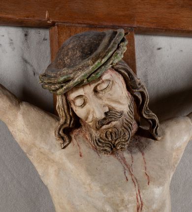 Zdjęcie nr 12: Rzeźba Chrystusa przybita do krzyża trzema gwoździami, pełna. Zbawiciel ukazany w typie Cristo morte, w lekkim zwisie, z głową pochyloną na prawą stronę. Jezus ma szczupłą sylwetkę ciała, duże stopy, delikatnie podkreśloną linię żeber. Jego twarz jest podłużna, ma długi nos, delikatnie otwarte usta i zamknięte oczy, krótką brodę, zwiniętą na końcu w dwa pukle. Ma długie, brązowe włosy spływające falami do przodu, a na głowie założoną koronę cierniową złożoną z prostych zielonych gałązek. Biodra oplata złocone perizonium z zieloną podszewką, zawiązane pośrodku. Z dłoni, stóp i boku Chrystusa spływają strużki krwi. Polichromia ciała naturalistyczna, perizonium złocone. Nad głową Jezusa titulus w formie banderoli z napisem „I. N. R. I.”. Krzyż prosty, gładki, malowany na brązowo.
