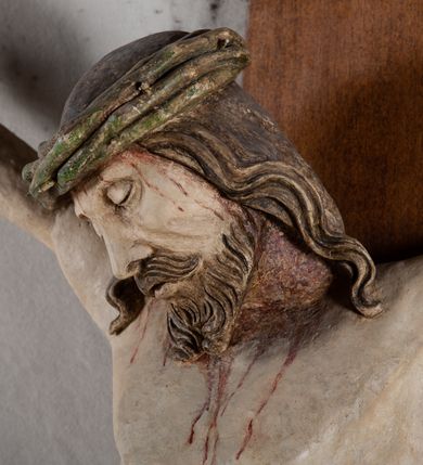 Zdjęcie nr 4: Rzeźba Chrystusa przybita do krzyża trzema gwoździami, pełna. Zbawiciel ukazany w typie Cristo morte, w lekkim zwisie, z głową pochyloną na prawą stronę. Jezus ma szczupłą sylwetkę ciała, duże stopy, delikatnie podkreśloną linię żeber. Jego twarz jest podłużna, ma długi nos, delikatnie otwarte usta i zamknięte oczy, krótką brodę, zwiniętą na końcu w dwa pukle. Ma długie, brązowe włosy spływające falami do przodu, a na głowie założoną koronę cierniową złożoną z prostych zielonych gałązek. Biodra oplata złocone perizonium z zieloną podszewką, zawiązane pośrodku. Z dłoni, stóp i boku Chrystusa spływają strużki krwi. Polichromia ciała naturalistyczna, perizonium złocone. Nad głową Jezusa titulus w formie banderoli z napisem „I. N. R. I.”. Krzyż prosty, gładki, malowany na brązowo.
