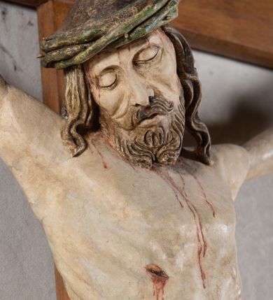 Zdjęcie nr 8: Rzeźba Chrystusa przybita do krzyża trzema gwoździami, pełna. Zbawiciel ukazany w typie Cristo morte, w lekkim zwisie, z głową pochyloną na prawą stronę. Jezus ma szczupłą sylwetkę ciała, duże stopy, delikatnie podkreśloną linię żeber. Jego twarz jest podłużna, ma długi nos, delikatnie otwarte usta i zamknięte oczy, krótką brodę, zwiniętą na końcu w dwa pukle. Ma długie, brązowe włosy spływające falami do przodu, a na głowie założoną koronę cierniową złożoną z prostych zielonych gałązek. Biodra oplata złocone perizonium z zieloną podszewką, zawiązane pośrodku. Z dłoni, stóp i boku Chrystusa spływają strużki krwi. Polichromia ciała naturalistyczna, perizonium złocone. Nad głową Jezusa titulus w formie banderoli z napisem „I. N. R. I.”. Krzyż prosty, gładki, malowany na brązowo.
