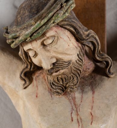 Zdjęcie nr 7: Rzeźba Chrystusa przybita do krzyża trzema gwoździami, pełna. Zbawiciel ukazany w typie Cristo morte, w lekkim zwisie, z głową pochyloną na prawą stronę. Jezus ma szczupłą sylwetkę ciała, duże stopy, delikatnie podkreśloną linię żeber. Jego twarz jest podłużna, ma długi nos, delikatnie otwarte usta i zamknięte oczy, krótką brodę, zwiniętą na końcu w dwa pukle. Ma długie, brązowe włosy spływające falami do przodu, a na głowie założoną koronę cierniową złożoną z prostych zielonych gałązek. Biodra oplata złocone perizonium z zieloną podszewką, zawiązane pośrodku. Z dłoni, stóp i boku Chrystusa spływają strużki krwi. Polichromia ciała naturalistyczna, perizonium złocone. Nad głową Jezusa titulus w formie banderoli z napisem „I. N. R. I.”. Krzyż prosty, gładki, malowany na brązowo.
