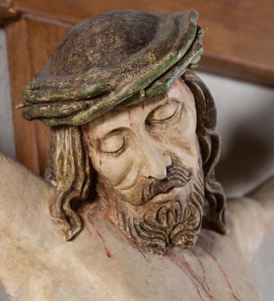 Zdjęcie nr 10: Rzeźba Chrystusa przybita do krzyża trzema gwoździami, pełna. Zbawiciel ukazany w typie Cristo morte, w lekkim zwisie, z głową pochyloną na prawą stronę. Jezus ma szczupłą sylwetkę ciała, duże stopy, delikatnie podkreśloną linię żeber. Jego twarz jest podłużna, ma długi nos, delikatnie otwarte usta i zamknięte oczy, krótką brodę, zwiniętą na końcu w dwa pukle. Ma długie, brązowe włosy spływające falami do przodu, a na głowie założoną koronę cierniową złożoną z prostych zielonych gałązek. Biodra oplata złocone perizonium z zieloną podszewką, zawiązane pośrodku. Z dłoni, stóp i boku Chrystusa spływają strużki krwi. Polichromia ciała naturalistyczna, perizonium złocone. Nad głową Jezusa titulus w formie banderoli z napisem „I. N. R. I.”. Krzyż prosty, gładki, malowany na brązowo.
