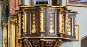 Zdjęcie nr 1: Ambona wisząca, przyścienna, z wejściem prowadzącym od zakrystii. 
Kosz na planie sześcioboku z podwieszeniem w postaci sześciobocznego stożka zakończonego szyszką. Ścianki kosza flankowane pilastrami podtrzymującymi niepełne belkowanie, zamknięte gzymsem wyłamanym na osi podpór tworzące parapet ambony. Ścianki podstawy dekorowane fantazyjną wicią roślinną złożoną ze stylizowanych liści akantu i kampanul. Ścianki kosza ozdobione prostokątnymi płycinami w złoconych, profilowanych ramach; w polu dekorowane stylizowaną, pionową wicią utworzoną z kampanul wyrastających z muszli. Pilastry ustawione na niskich cokolikach zdobionych rozetami, z trzonami z szerokim pasem utworzonym z liści o ostrych zakończeniach. 

W zaplecku, w prostokątnej ramie współczesny obraz Jezusa Dobrego Pasterza, namalowany  przez Zbigniewa Juszczaka. 
Baldachim na planie sześcioboku od dołu zdobiony lambrekinem, zamknięty od góry profilowanym gzymsem, w zwieńczeniu z bogato dekorowanym krzyżem łacińskim stojącym na ostrosłupie. U spodu baldachimu gołębica Ducha Świętego na tle promieni ułożonych w wiązki o nierównej długości. 
Struktura polichromowana na kolor ciemnobrązowy, ornamentyka i profilowania złocone.
