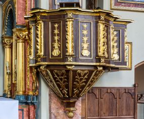 Zdjęcie nr 1: Ambona wisząca, przyścienna, z wejściem prowadzącym od zakrystii. 
Kosz na planie sześcioboku z podwieszeniem w postaci sześciobocznego stożka zakończonego szyszką. Ścianki kosza flankowane pilastrami podtrzymującymi niepełne belkowanie, zamknięte gzymsem wyłamanym na osi podpór tworzące parapet ambony. Ścianki podstawy dekorowane fantazyjną wicią roślinną złożoną ze stylizowanych liści akantu i kampanul. Ścianki kosza ozdobione prostokątnymi płycinami w złoconych, profilowanych ramach; w polu dekorowane stylizowaną, pionową wicią utworzoną z kampanul wyrastających z muszli. Pilastry ustawione na niskich cokolikach zdobionych rozetami, z trzonami z szerokim pasem utworzonym z liści o ostrych zakończeniach. 

W zaplecku, w prostokątnej ramie współczesny obraz Jezusa Dobrego Pasterza, namalowany  przez Zbigniewa Juszczaka. 
Baldachim na planie sześcioboku od dołu zdobiony lambrekinem, zamknięty od góry profilowanym gzymsem, w zwieńczeniu z bogato dekorowanym krzyżem łacińskim stojącym na ostrosłupie. U spodu baldachimu gołębica Ducha Świętego na tle promieni ułożonych w wiązki o nierównej długości. 
Struktura polichromowana na kolor ciemnobrązowy, ornamentyka i profilowania złocone.
