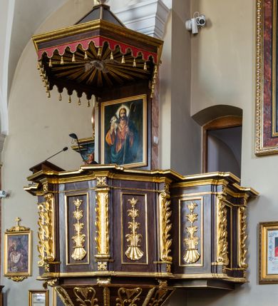 Zdjęcie nr 2: Ambona wisząca, przyścienna, z wejściem prowadzącym od zakrystii. 
Kosz na planie sześcioboku z podwieszeniem w postaci sześciobocznego stożka zakończonego szyszką. Ścianki kosza flankowane pilastrami podtrzymującymi niepełne belkowanie, zamknięte gzymsem wyłamanym na osi podpór tworzące parapet ambony. Ścianki podstawy dekorowane fantazyjną wicią roślinną złożoną ze stylizowanych liści akantu i kampanul. Ścianki kosza ozdobione prostokątnymi płycinami w złoconych, profilowanych ramach; w polu dekorowane stylizowaną, pionową wicią utworzoną z kampanul wyrastających z muszli. Pilastry ustawione na niskich cokolikach zdobionych rozetami, z trzonami z szerokim pasem utworzonym z liści o ostrych zakończeniach. 

W zaplecku, w prostokątnej ramie współczesny obraz Jezusa Dobrego Pasterza, namalowany  przez Zbigniewa Juszczaka. 
Baldachim na planie sześcioboku od dołu zdobiony lambrekinem, zamknięty od góry profilowanym gzymsem, w zwieńczeniu z bogato dekorowanym krzyżem łacińskim stojącym na ostrosłupie. U spodu baldachimu gołębica Ducha Świętego na tle promieni ułożonych w wiązki o nierównej długości. 
Struktura polichromowana na kolor ciemnobrązowy, ornamentyka i profilowania złocone.
