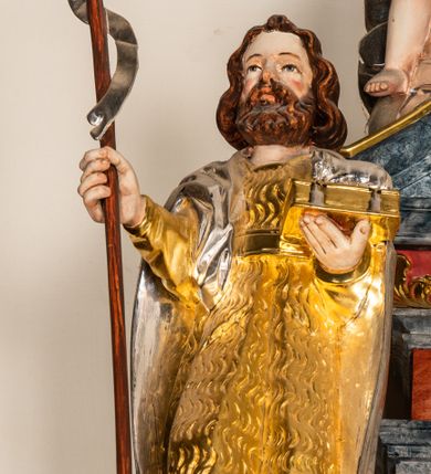 Zdjęcie nr 1: Drążona z tyłu, całopostaciowa figura św. Jana Chrzciciela stojącego frontalnie. Głowa lekko w trzech czwartych zwrócona w prawo, delikatnie odchylona do tyłu. Twarz owalna o wyrazistych rysach, z brązowym, krótkim, kręconym zarostem. Włosy brązowe, półdługie, mocno falowane, z przedziałkiem pośrodku. Święty ubrany w sięgającą kolan, złoconą skórę przewiązaną w pasie, na ramiona ma narzucony srebrzony, sięgający kolan płaszcz. W prawej dłoni trzyma brązowy krzyż na długim drzewcu, ze srebrzoną wąską szarfą. W lewej dłoni podtrzymuje zamkniętą, złoconą księgę z leżącym na niej srebrzonym barankiem. 