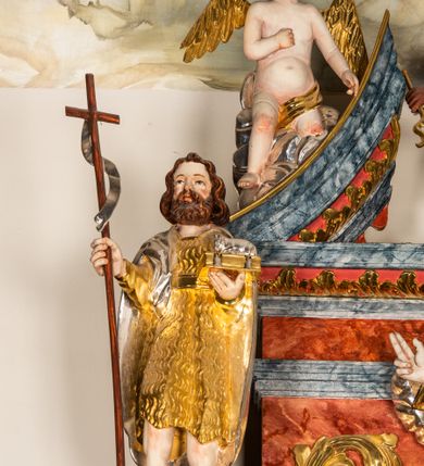 Zdjęcie nr 2: Drążona z tyłu, całopostaciowa figura św. Jana Chrzciciela stojącego frontalnie. Głowa lekko w trzech czwartych zwrócona w prawo, delikatnie odchylona do tyłu. Twarz owalna o wyrazistych rysach, z brązowym, krótkim, kręconym zarostem. Włosy brązowe, półdługie, mocno falowane, z przedziałkiem pośrodku. Święty ubrany w sięgającą kolan, złoconą skórę przewiązaną w pasie, na ramiona ma narzucony srebrzony, sięgający kolan płaszcz. W prawej dłoni trzyma brązowy krzyż na długim drzewcu, ze srebrzoną wąską szarfą. W lewej dłoni podtrzymuje zamkniętą, złoconą księgę z leżącym na niej srebrzonym barankiem. 