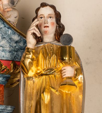 Zdjęcie nr 1: Drążona z tyłu, całopostaciowa figura św. Jana Ewangelisty stojącego frontalnie w lekkim kontrapoście. Twarz owalna o delikatnych rysach. Włosy brązowe, długie, mocno falowane. Święty jest ubrany w długą, złoconą suknię przewiązaną w pasie, na ramiona ma narzucony srebrzony, długi płaszcz spięty pod szyją. Prawa ręka w geście błogosławieństwa, w lewej dłoni złocony kielich ze srebrną hostią.  