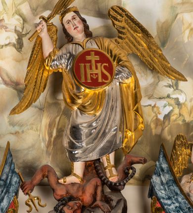 Zdjęcie nr 1: Ścięta z tyłu, całopostaciowa figura św. Michała Archanioła stojącego frontalnie w lekkim kontrapoście, o sylwetce lekko przechylonej w prawo, pokonującego szatana. Twarz owalna o delikatnych rysach. Włosy brązowe, długie, falowane, opadające na plecy. Na głowie złocony diadem. Archanioł ubrany jest w długą, srebrzoną suknię i przerzucony przez lewe ramię złocony, podtrzymywany lewą ręką płaszcz. Na stopach złocone sandały. W uniesionej prawej ręce miecz ognisty. W lewej, osłaniająca tułów okrągła tarcza ze złoconym monogramem chrystologicznym na czerwonym tle. Na plecach złocone skrzydła. Pod stopami postać leżącego na skale szatana o ciemnobrązowej karnacji, z krótkimi, kręconymi, czarnymi włosami, z długim ogonem. W lewej dłoni złocony trójząb.   