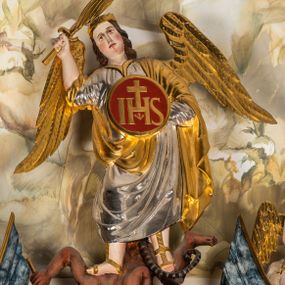 Zdjęcie nr 1: Ścięta z tyłu, całopostaciowa figura św. Michała Archanioła stojącego frontalnie w lekkim kontrapoście, o sylwetce lekko przechylonej w prawo, pokonującego szatana. Twarz owalna o delikatnych rysach. Włosy brązowe, długie, falowane, opadające na plecy. Na głowie złocony diadem. Archanioł ubrany jest w długą, srebrzoną suknię i przerzucony przez lewe ramię złocony, podtrzymywany lewą ręką płaszcz. Na stopach złocone sandały. W uniesionej prawej ręce miecz ognisty. W lewej, osłaniająca tułów okrągła tarcza ze złoconym monogramem chrystologicznym na czerwonym tle. Na plecach złocone skrzydła. Pod stopami postać leżącego na skale szatana o ciemnobrązowej karnacji, z krótkimi, kręconymi, czarnymi włosami, z długim ogonem. W lewej dłoni złocony trójząb.   