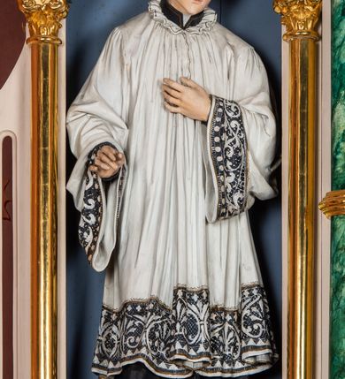 Zdjęcie nr 13: Ołtarz przyścienny, architektoniczny, jednokondygnacyjny, trójosiowy, ze zwieńczeniem. Ołtarz prostopadłościenny z antepedium artykułowanym trzema płycinami. Dwie skrajne, węższe zawierają dekorację złożoną z korony i nasadzonego na nią krzyża pomiędzy liśćmi wawrzynu ustawionymi w słup, środkowa, szersza z motywem krzyża równoramiennego o trójlistnych zakończeniach ramion, ujętego maswerkiem. Predella wklęśle zwężająca się, po bokach artykułowana pięcioma płycinami ze stylizowanymi wiciami roślinnymi. Tabernakulum w formie prostopadłościennej szafki, flankowane od frontu parą kolumienek wtopionych w naroża, zamknięte nadwieszonym łukiem półkolistym. Prostokątne, półkoliście zamknięte drzwiczki zdobione płaskorzeźbionym motywem kielicha eucharystycznego.  Część środkowa nastawy w formie trójprzelotowego łuku triumfalnego. Każda z ujmujących płytkie wnęki półkolistych arkad wsparta na parze kolumn. Podłucza ujęte dekoracyjnym ornamentem arkadkowym, który w części środkowej przybiera kształt trójliścia. Kolumny wspierają zamykające osie wimpergi. Zwieńczenie części środkowej retabulum osadzone na zdwojonym cokole, ma formę wspartej na parze kolumienek półkolistej arkady, której podłucze wypełnia motyw trójliścia, zamkniętej wimpergą. Całość ujmuje para przypór o zakończeniach ujętych kolumienkami wspierającymi sterczyny. Boczne osie zwieńczone motywem oktogonalnych latarni przekrytych kopułkami. Pola predelli zdobią motywy wici akantowych, boki ujmuje para ażurowych uszaków. W polach wimperg oraz w dolnej partii wnęki części środkowej stylizowana ornamentyka roślinna, w górnej części monogram maryjny. Krawędzie zwieńczenia zdobi motyw wici roślinnej, czołganki oraz sterczyny zwieńczone kwiatonami. We wnęce części środkowej ujęty rzeźbioną, złoconą ramą obraz Matki Boskiej z Dzieciątkiem. We wnęce po prawej stronie retabulum figura św. Stanisława Kostki, po lewej św. Alojzego Gonzagi. W zwieńczeniu figura anioła. Polichromia ołtarza utrzymana w kolorach beżu i różu, detale i ornamenty złocone. Kolumny środkowej osi zielone, marmoryzowane, gzymsy w przyczółkach w kolorach niebieskim i czerwonym.