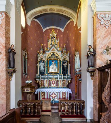 Zdjęcie nr 2: Ołtarz przyścienny, architektoniczny, jednokondygnacyjny, trójosiowy, ze zwieńczeniem. Ołtarz prostopadłościenny z antepedium artykułowanym trzema płycinami. Dwie skrajne, węższe zawierają dekorację złożoną z korony i nasadzonego na nią krzyża pomiędzy liśćmi wawrzynu ustawionymi w słup, środkowa, szersza z motywem krzyża równoramiennego o trójlistnych zakończeniach ramion, ujętego maswerkiem. Predella wklęśle zwężająca się, po bokach artykułowana pięcioma płycinami ze stylizowanymi wiciami roślinnymi. Tabernakulum w formie prostopadłościennej szafki, flankowane od frontu parą kolumienek wtopionych w naroża, zamknięte nadwieszonym łukiem półkolistym. Prostokątne, półkoliście zamknięte drzwiczki zdobione płaskorzeźbionym motywem kielicha eucharystycznego.  Część środkowa nastawy w formie trójprzelotowego łuku triumfalnego. Każda z ujmujących płytkie wnęki półkolistych arkad wsparta na parze kolumn. Podłucza ujęte dekoracyjnym ornamentem arkadkowym, który w części środkowej przybiera kształt trójliścia. Kolumny wspierają zamykające osie wimpergi. Zwieńczenie części środkowej retabulum osadzone na zdwojonym cokole, ma formę wspartej na parze kolumienek półkolistej arkady, której podłucze wypełnia motyw trójliścia, zamkniętej wimpergą. Całość ujmuje para przypór o zakończeniach ujętych kolumienkami wspierającymi sterczyny. Boczne osie zwieńczone motywem oktogonalnych latarni przekrytych kopułkami. Pola predelli zdobią motywy wici akantowych, boki ujmuje para ażurowych uszaków. W polach wimperg oraz w dolnej partii wnęki części środkowej stylizowana ornamentyka roślinna, w górnej części monogram maryjny. Krawędzie zwieńczenia zdobi motyw wici roślinnej, czołganki oraz sterczyny zwieńczone kwiatonami. We wnęce części środkowej ujęty rzeźbioną, złoconą ramą obraz Matki Boskiej z Dzieciątkiem. We wnęce po prawej stronie retabulum figura św. Stanisława Kostki, po lewej św. Alojzego Gonzagi. W zwieńczeniu figura anioła. Polichromia ołtarza utrzymana w kolorach beżu i różu, detale i ornamenty złocone. Kolumny środkowej osi zielone, marmoryzowane, gzymsy w przyczółkach w kolorach niebieskim i czerwonym.